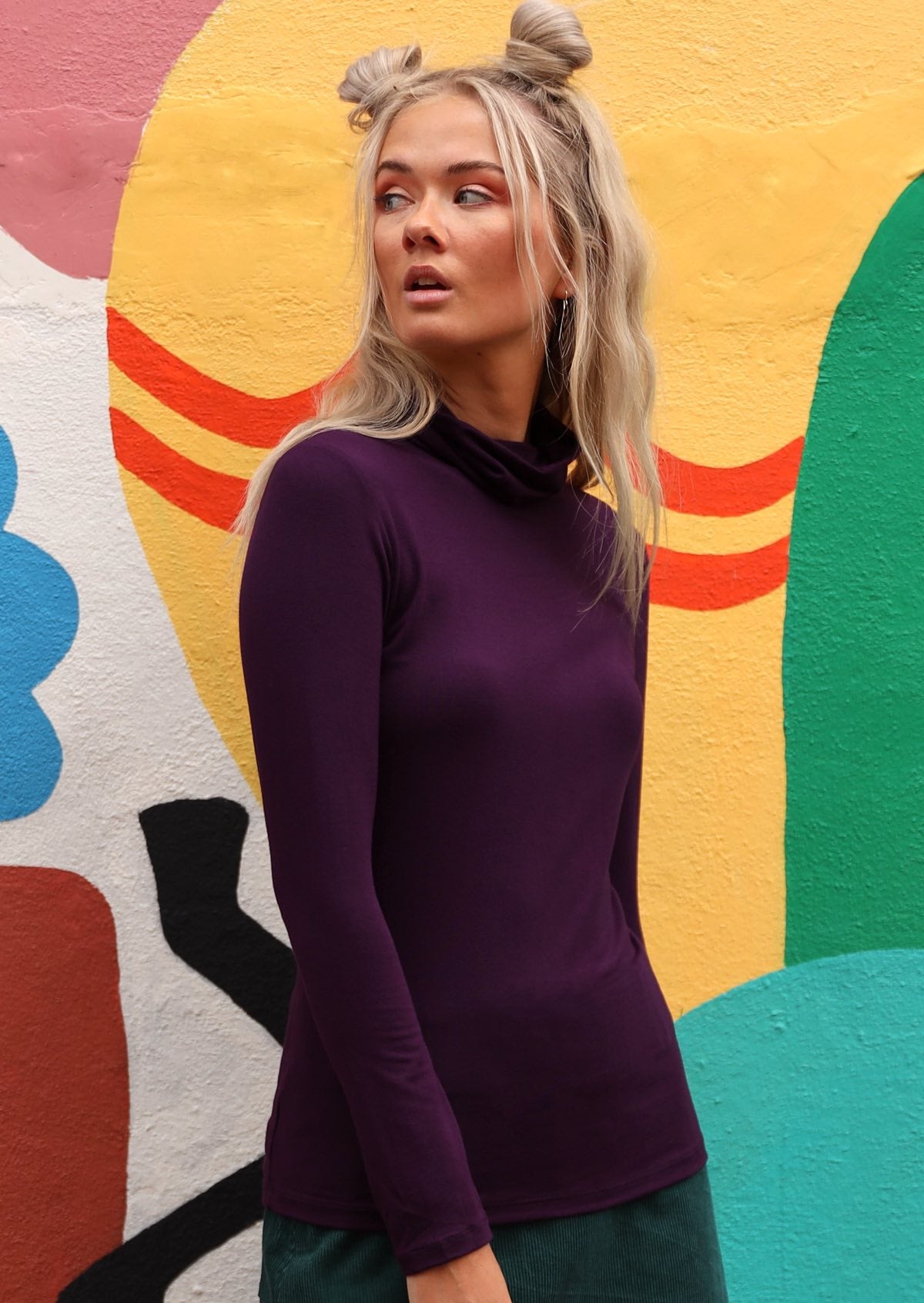 Woman wearing a turtle neck purple fitted long sleeve soft stretch rayon top in front of colourful wall.