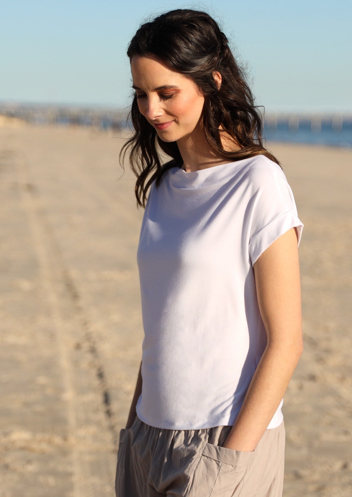 Woman with dark hair standing on the beach wearing a wide neck mod white stretch rayon boat neck top
