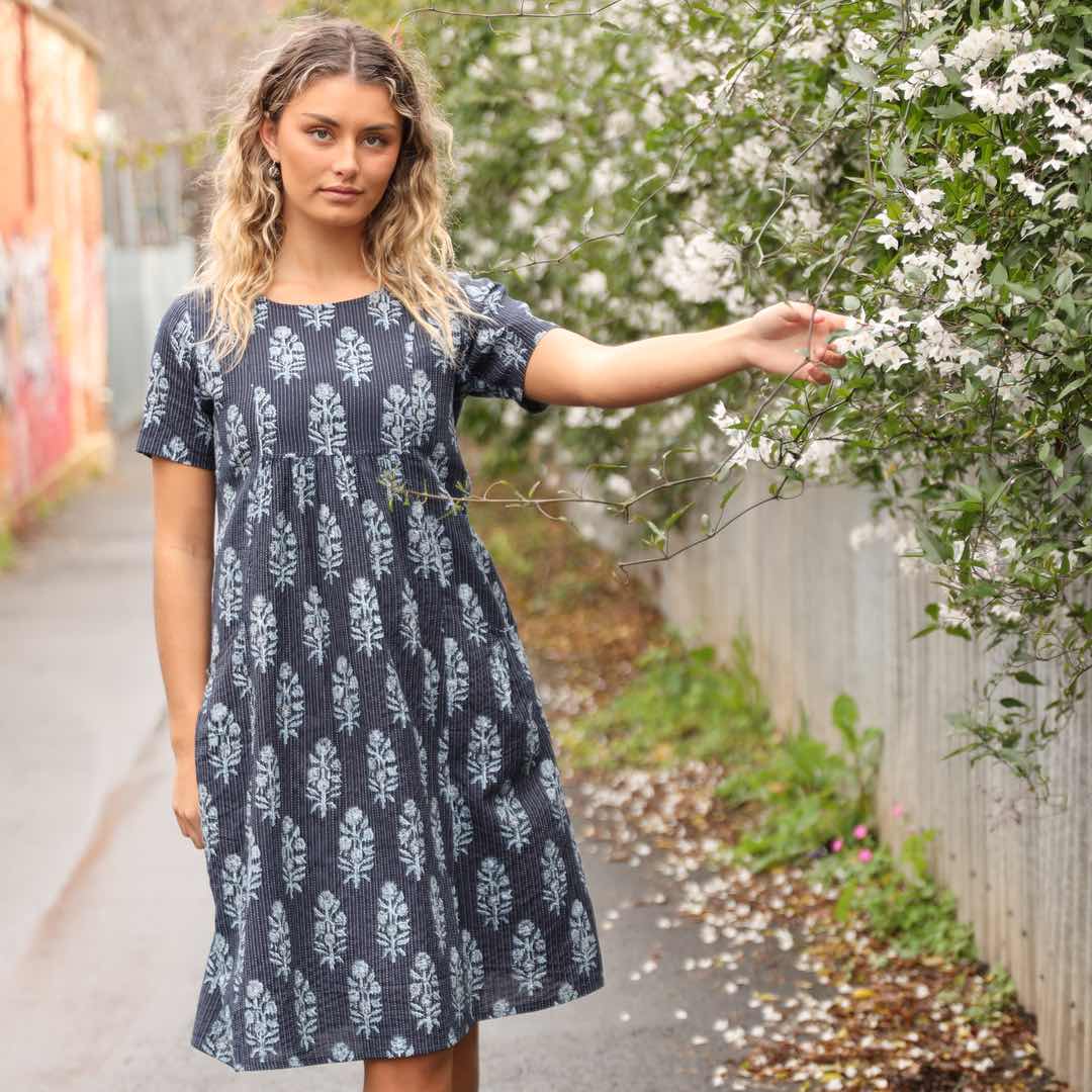 woman wearing navy blue cotton dress with Indian print in lane way with flowers cascading over fence