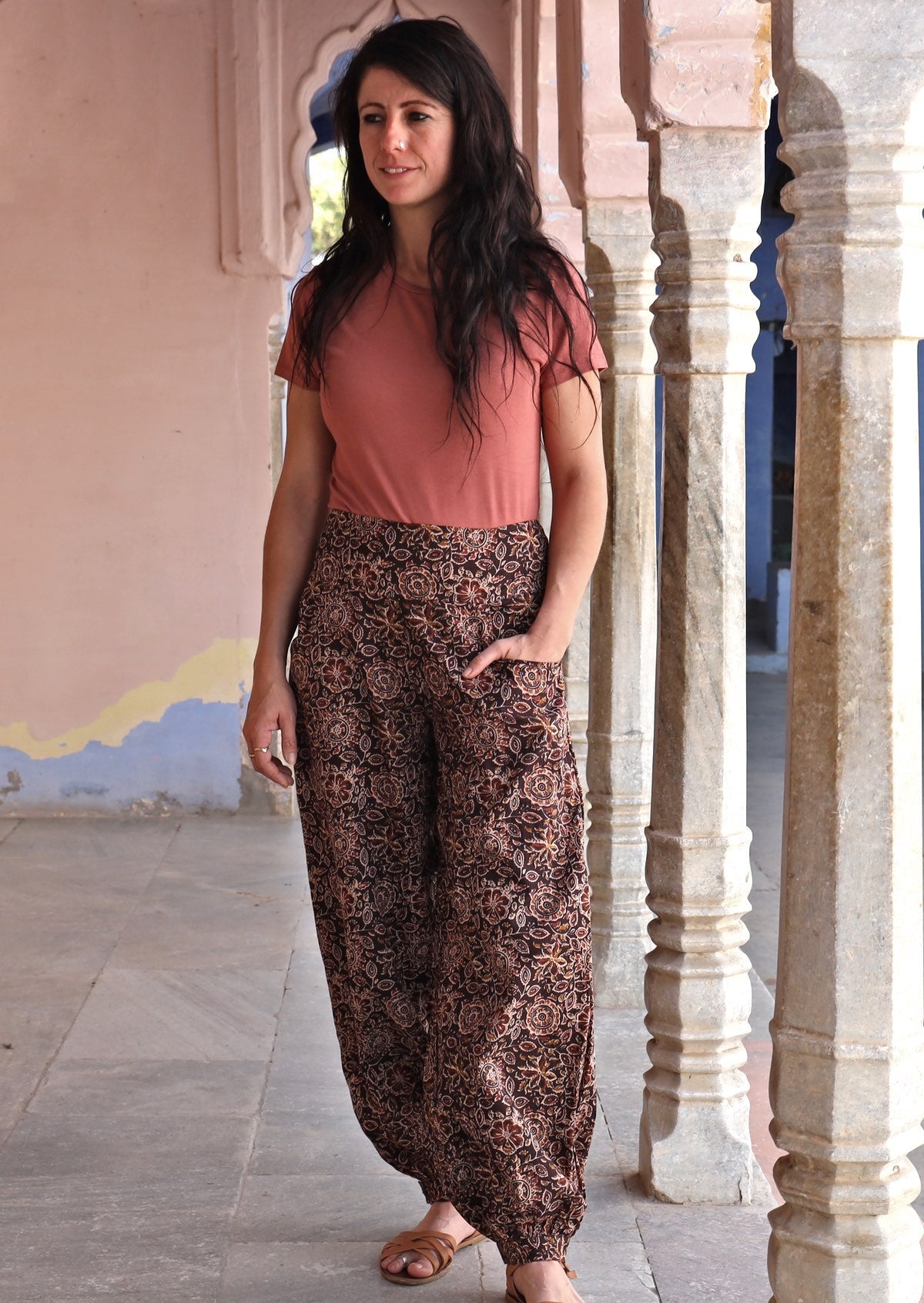 Model posing in floral printed cotton pants in brown with cuffed ankles and a hand in pocket.