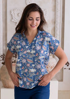 Woman wearing a blue based relaxed fit, shirt style floral cotton top with hands in pocket of dark blue cotton pants.