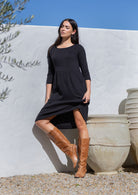 Woman in a black cotton gauze dress and brown boots standing against a white wall with blue sky.
