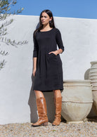 Woman wearing a black double cotton gauze dress and brown knee-high boots standing against a white wall with a blue sky.