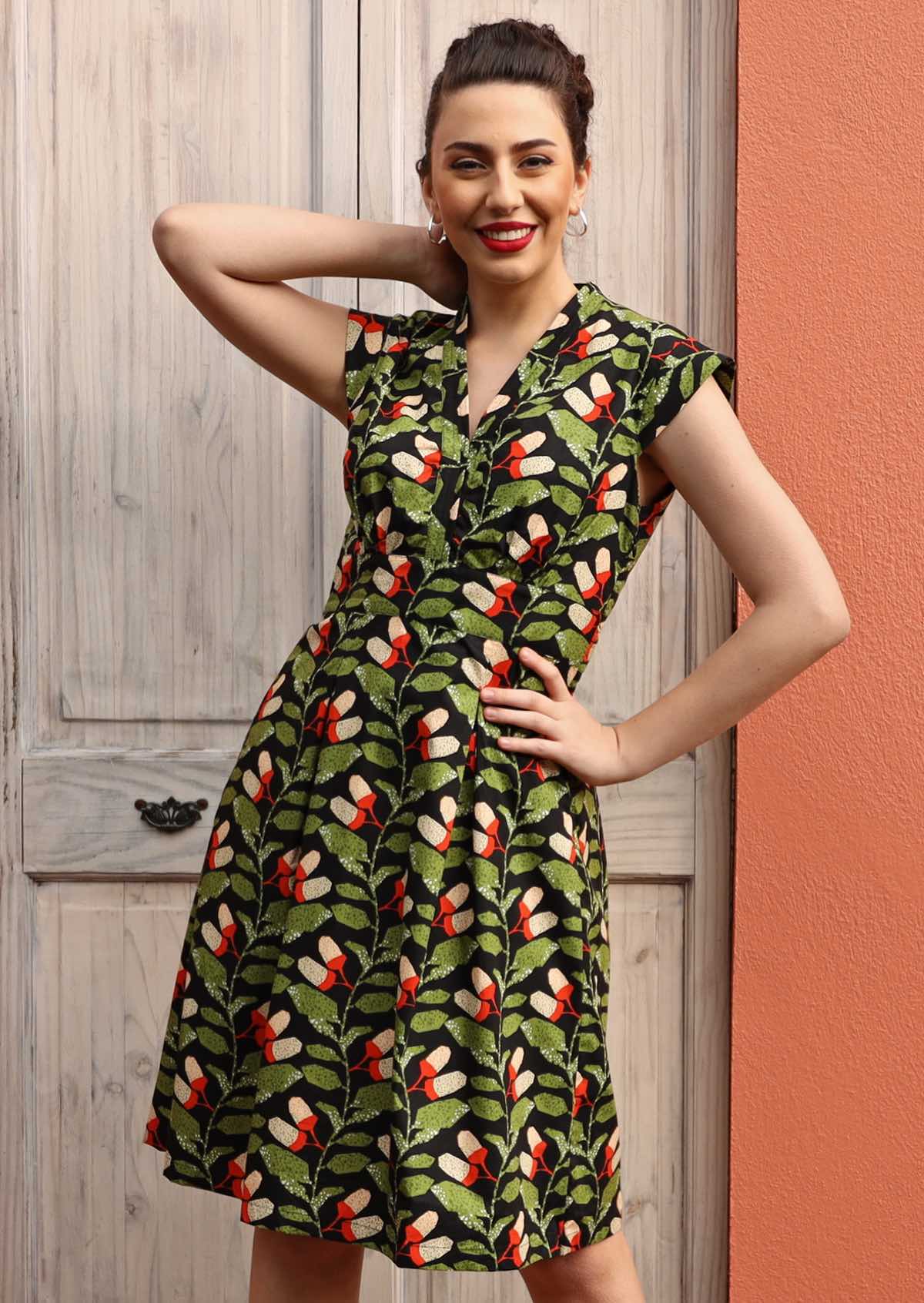 Model wears Billie Dress Oak green, orange- white speckled botanical print on black base cotton V-neck cap sleeves box pleats at waist form A-line skirt side zip knee length 