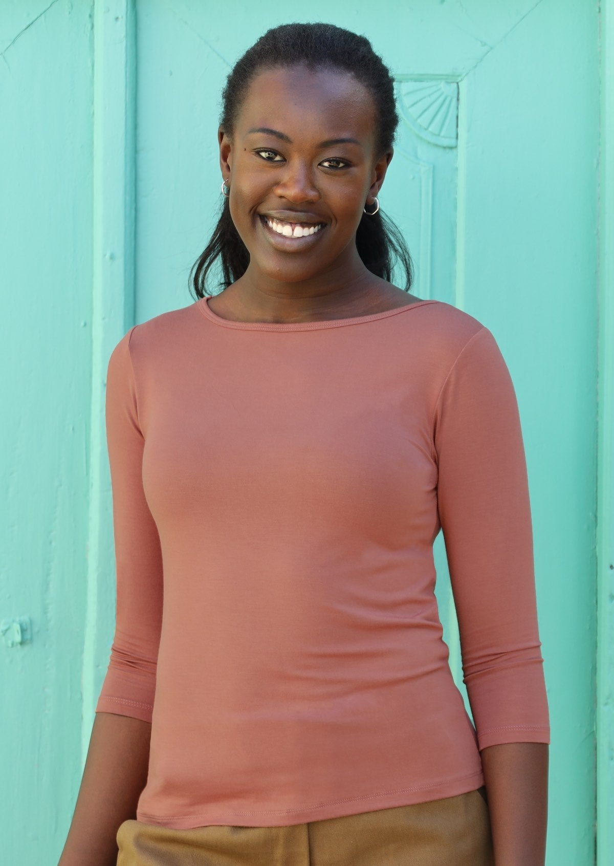 Woman wearing a rayon boat neck dusty pink 3/4 sleeve top in front of green wall.