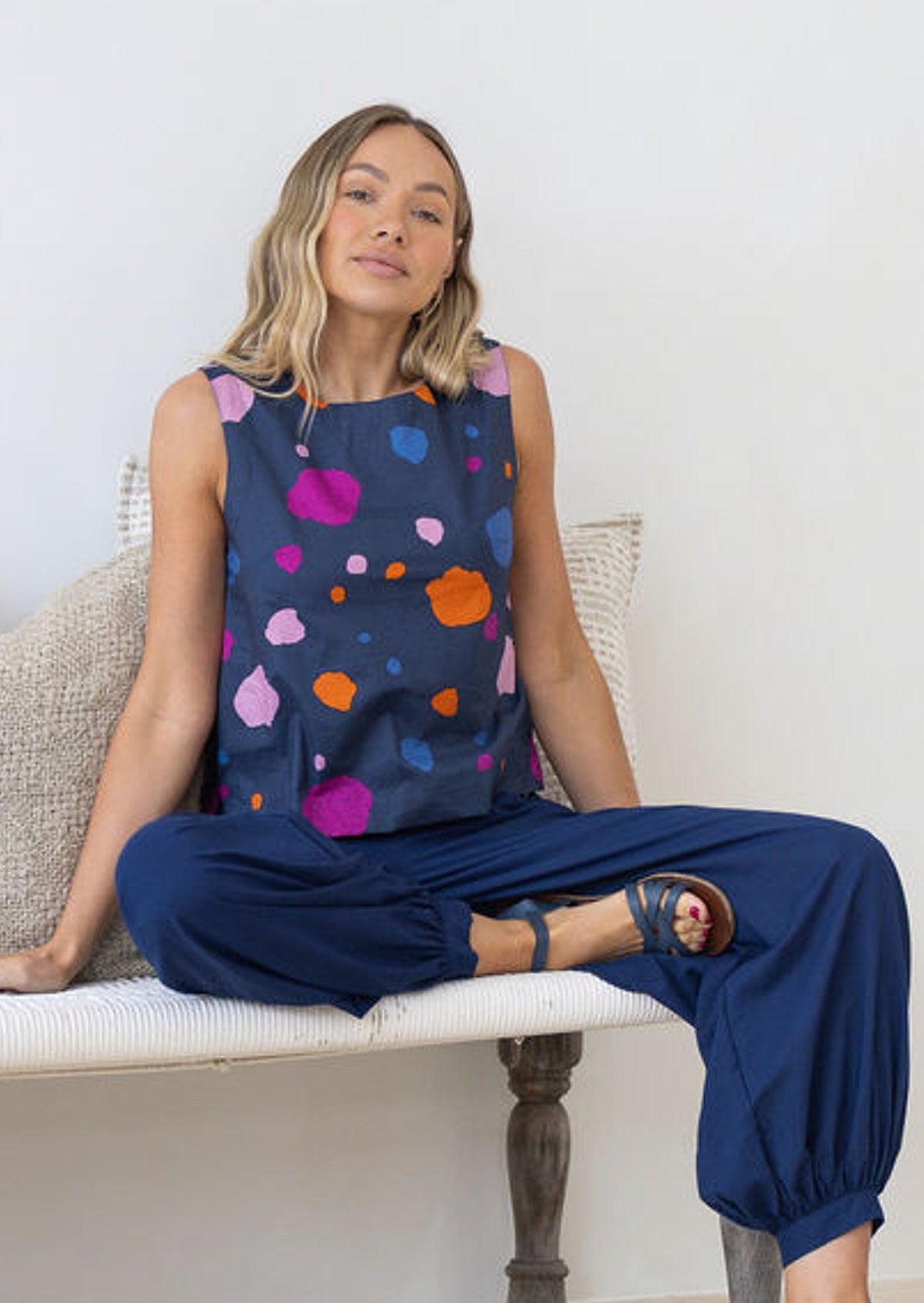 Woman sitting on a chair wearing a navy sleeveless top with colourful polka dots and navy blue pants.
