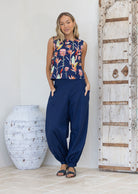 Woman wearing a sleeveless floral top and blue pants standing in a room with a large vase and wooden door.