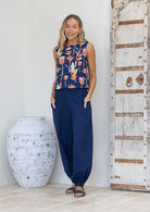 Woman wearing a navy sleeveless top with colourful floral pattern and navy pants, standing next to a large decorative vase and wooden door.