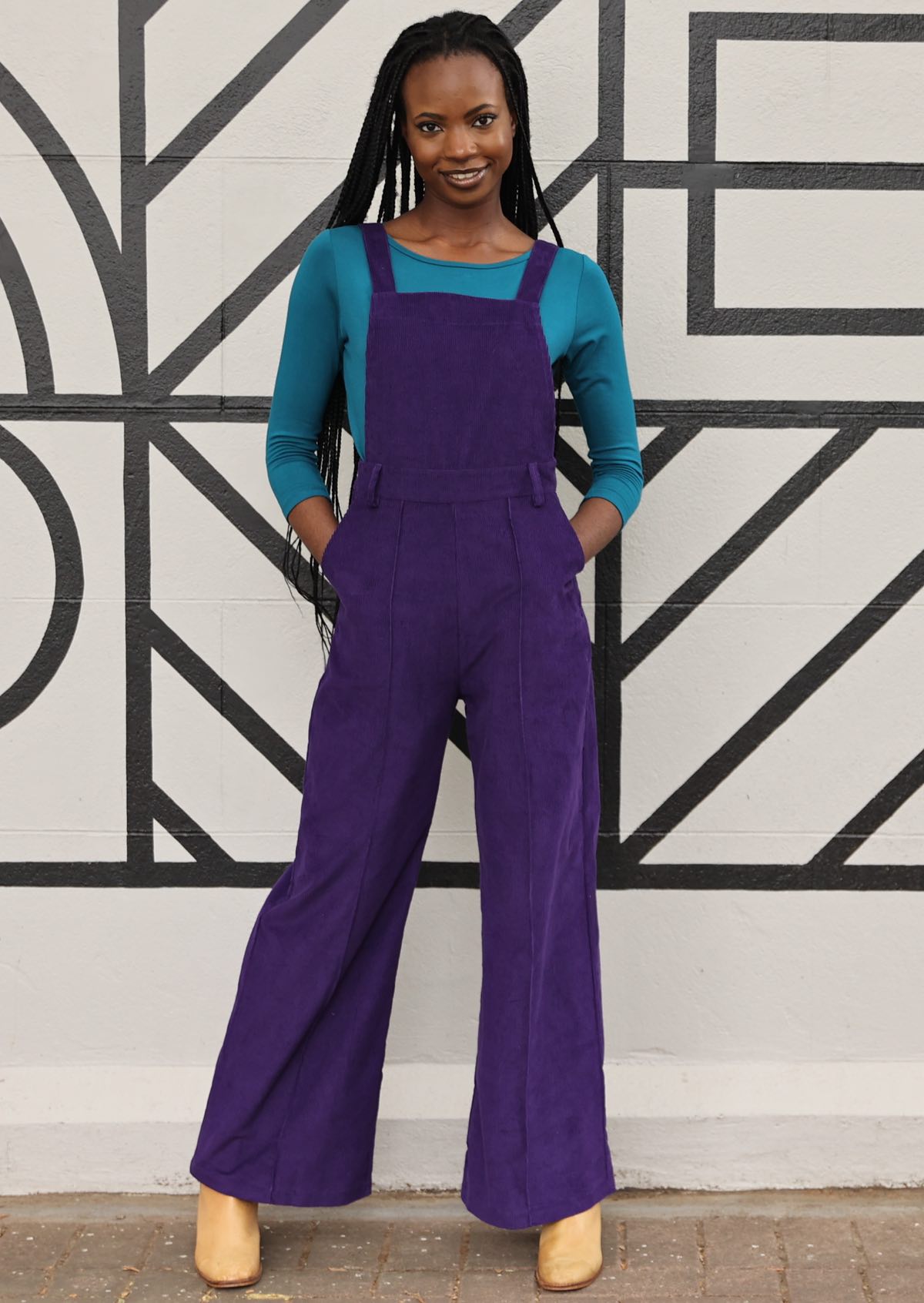 model wearing purple cotton corduroy overalls with hands in pockets
