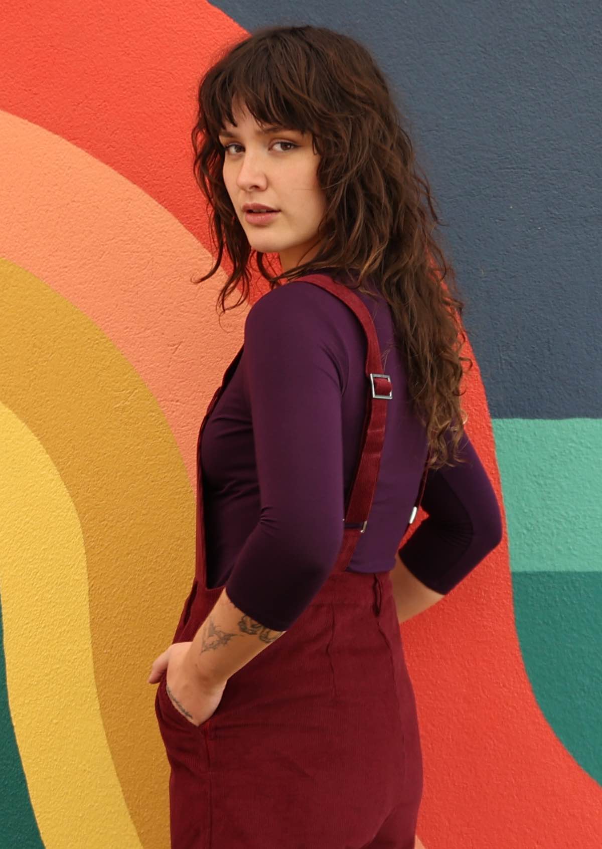 woman wearing maroon cotton corduroy overalls over long sleeved purple top back view