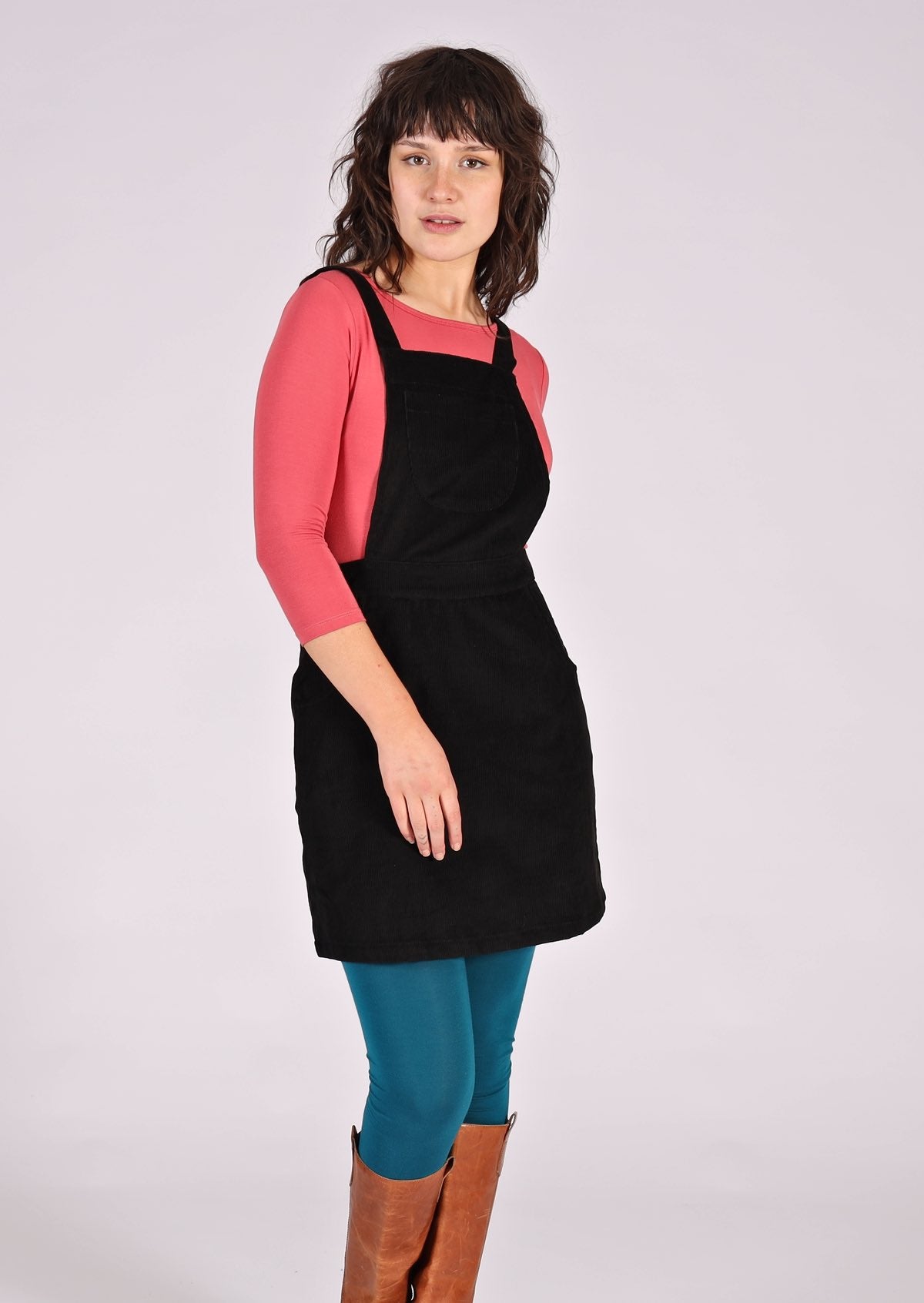 woman wearing black corduroy pinafore over pink top, teal leggings and tan boots