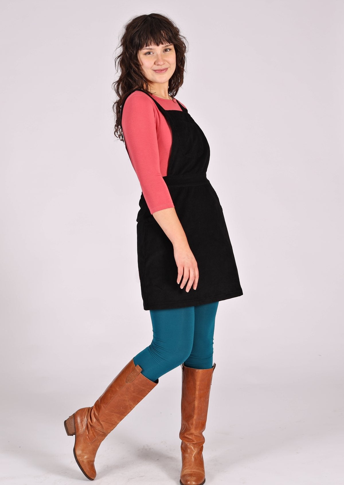 woman wearing black corduroy pinafore over pink top and teal leggings
