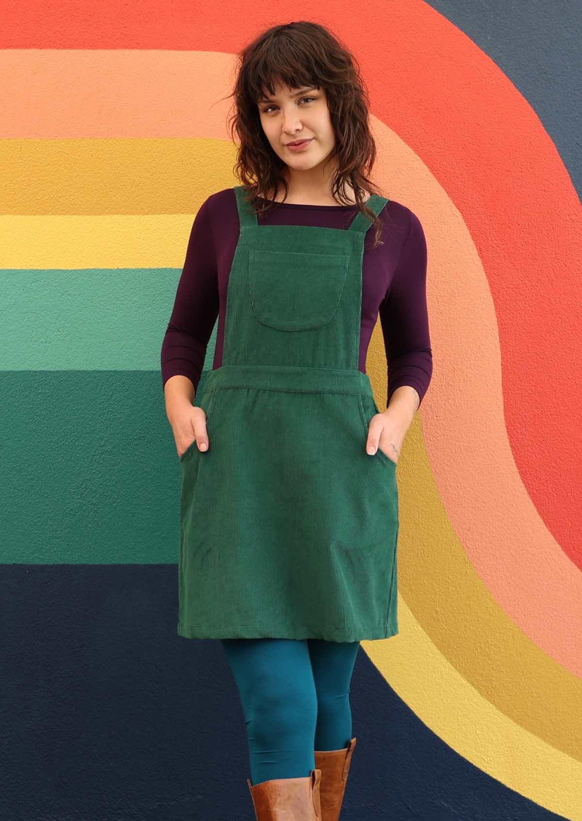woman wearing green corduroy pinafore over purple top and teal leggings both hands in pockets