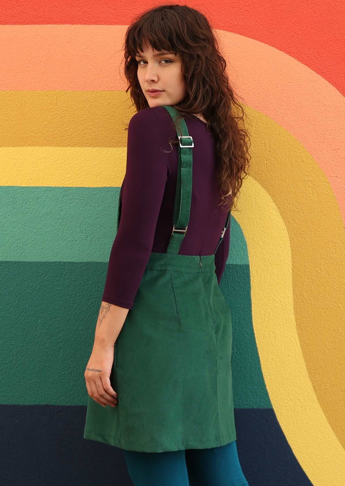 woman wearing green corduroy pinafore over purple top and teal leggings back view