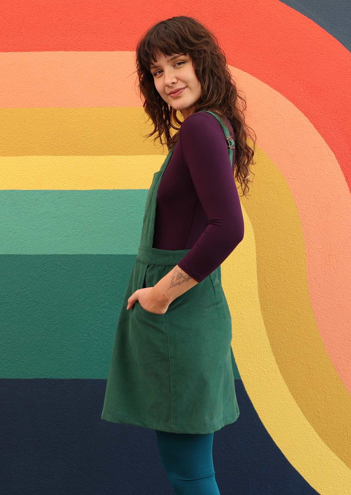 woman wearing green corduroy pinafore over purple top and teal leggings side view