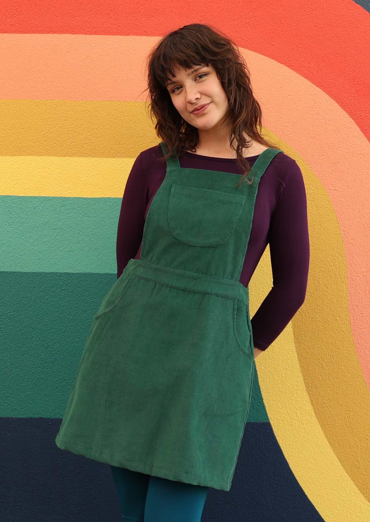 woman wearing green corduroy pinafore over purple top and teal leggings 