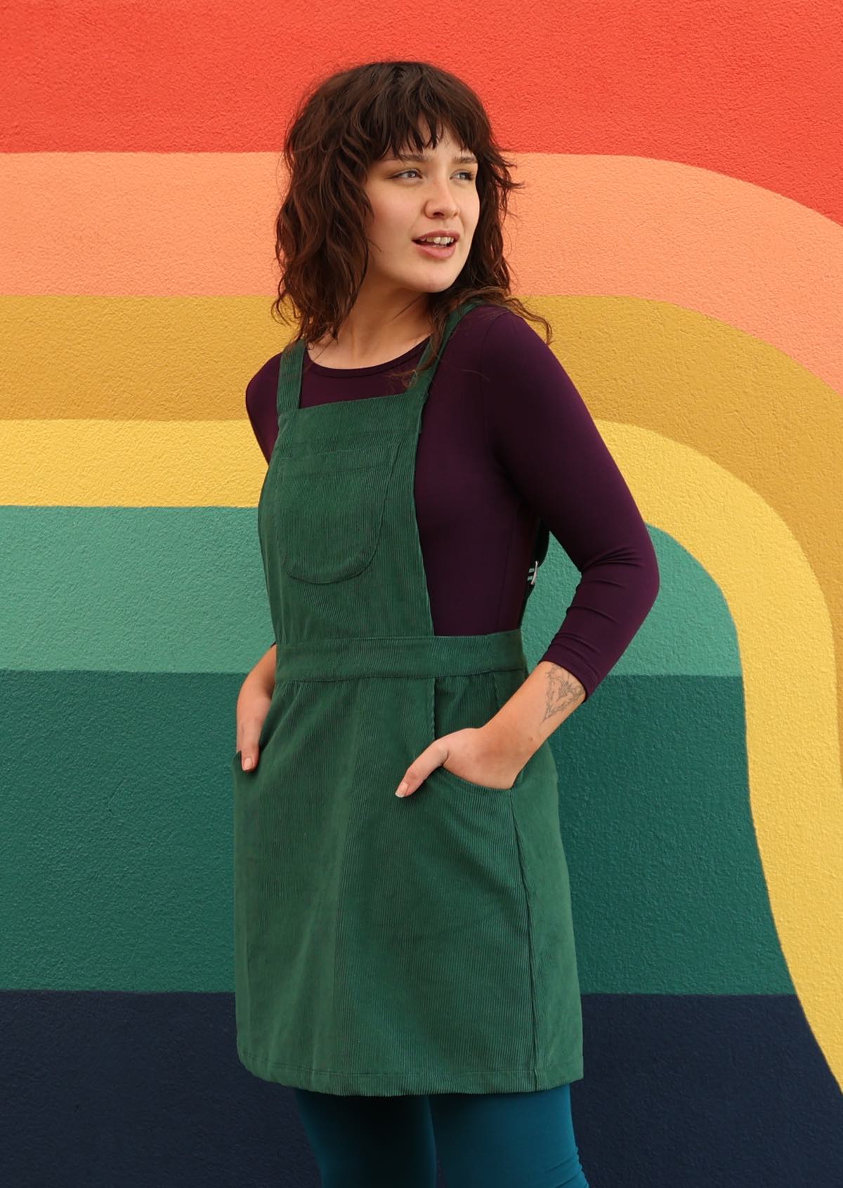 woman wearing green corduroy pinafore over purple top and teal leggings with hands in pockets