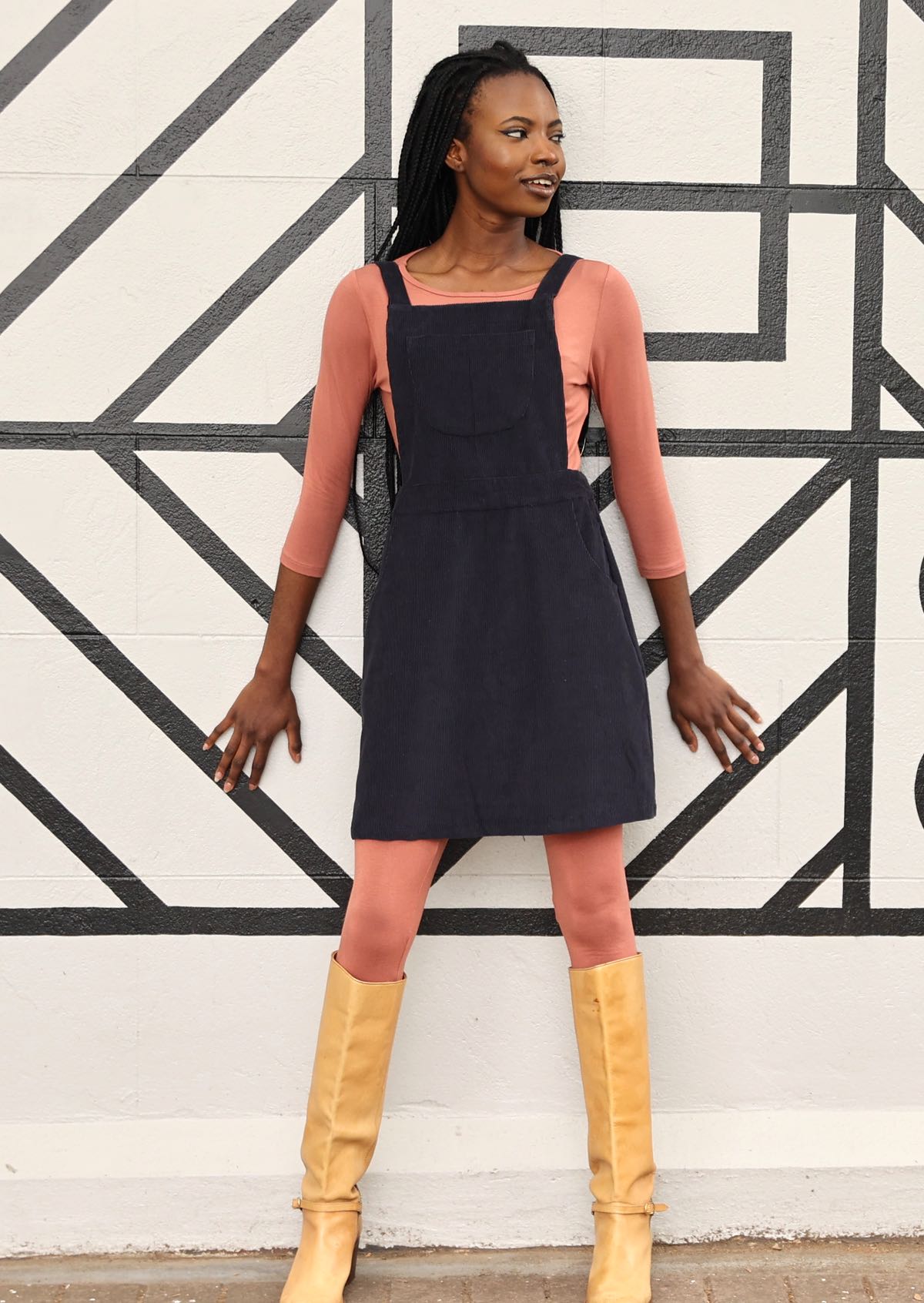 woman wearing dark grey cotton corduroy pinafore over pink top and pink leggings  with tan boots