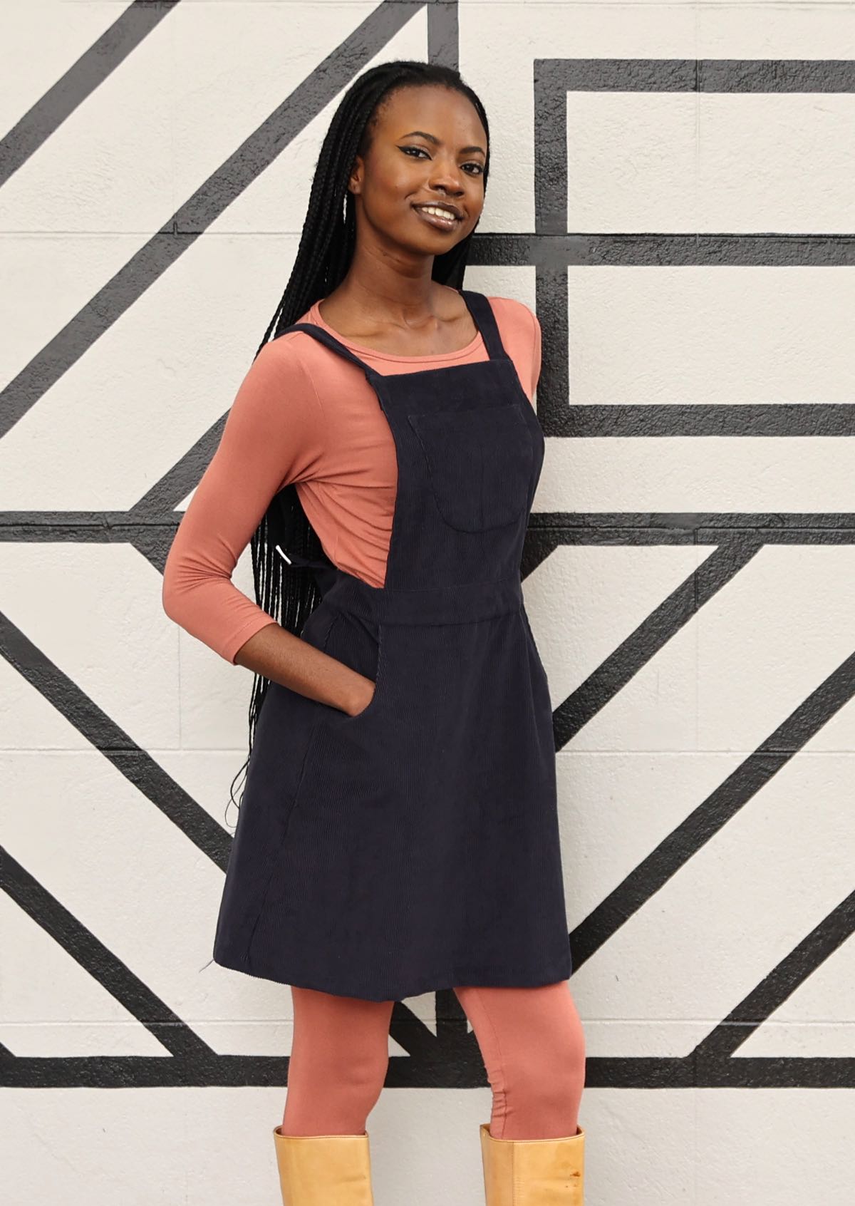 woman wearing dark grey cotton corduroy pinafore with pockets over pink top and pink leggings  