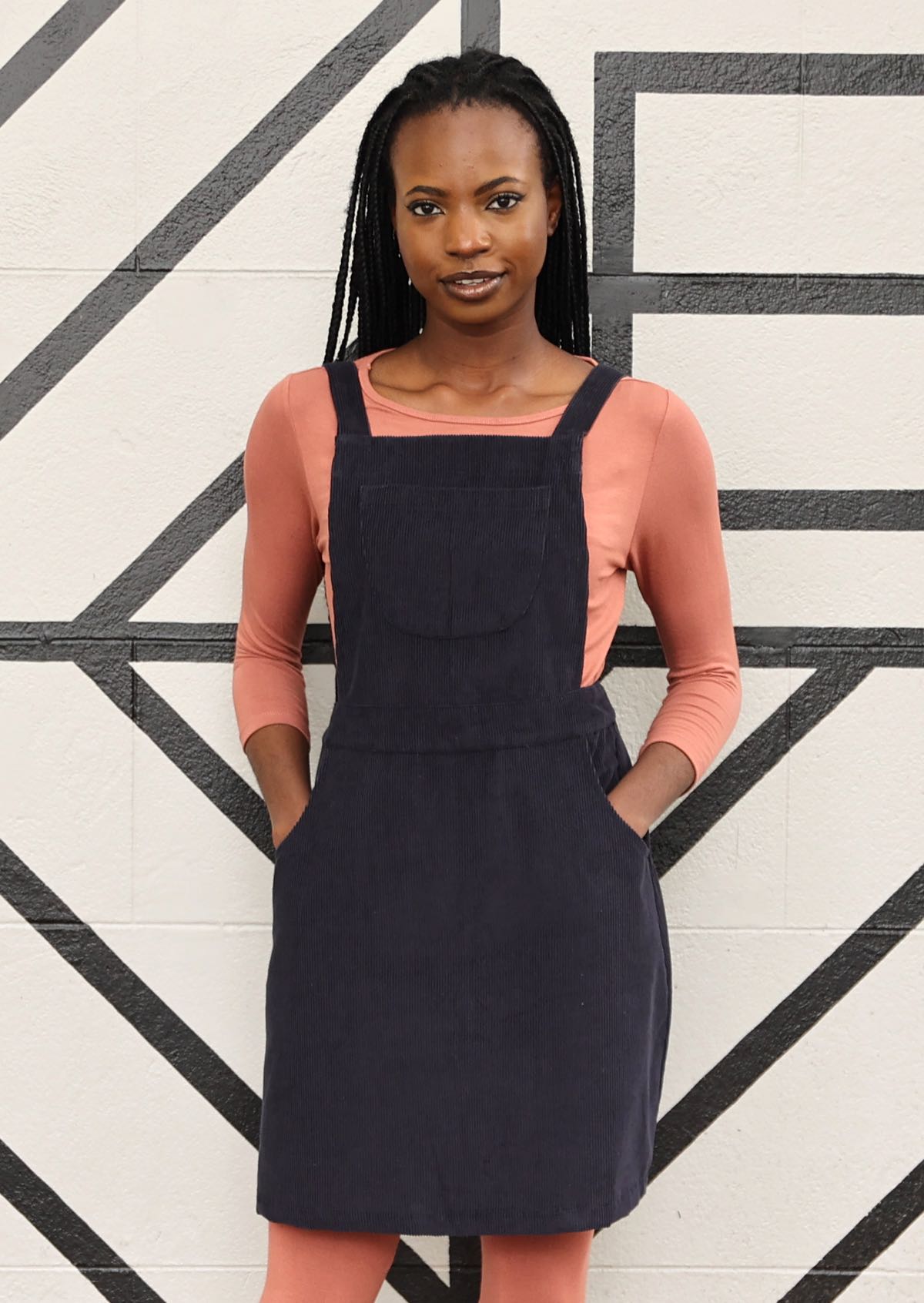 woman wearing dark grey cotton corduroy pinafore over pink top and pink leggings  with hands in pockets
