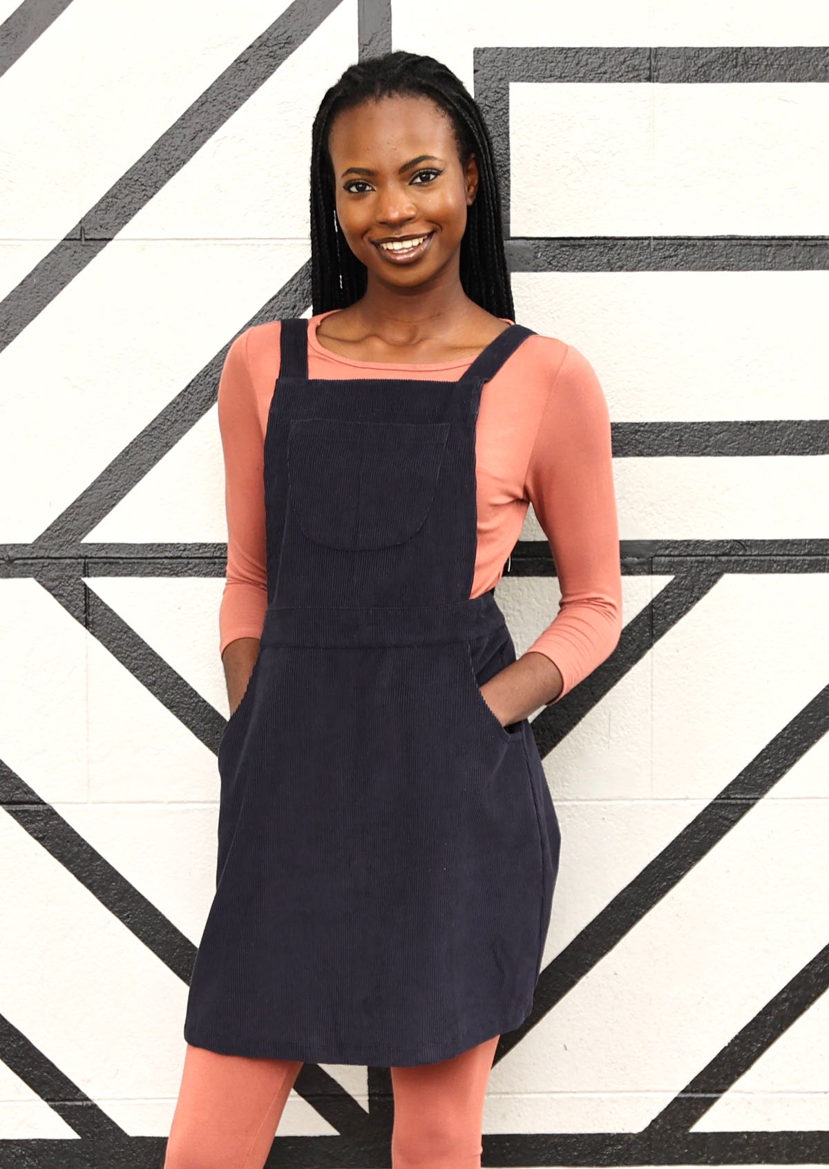 woman wearing dark grey cotton corduroy pinafore over pink top and soft pink leggings 