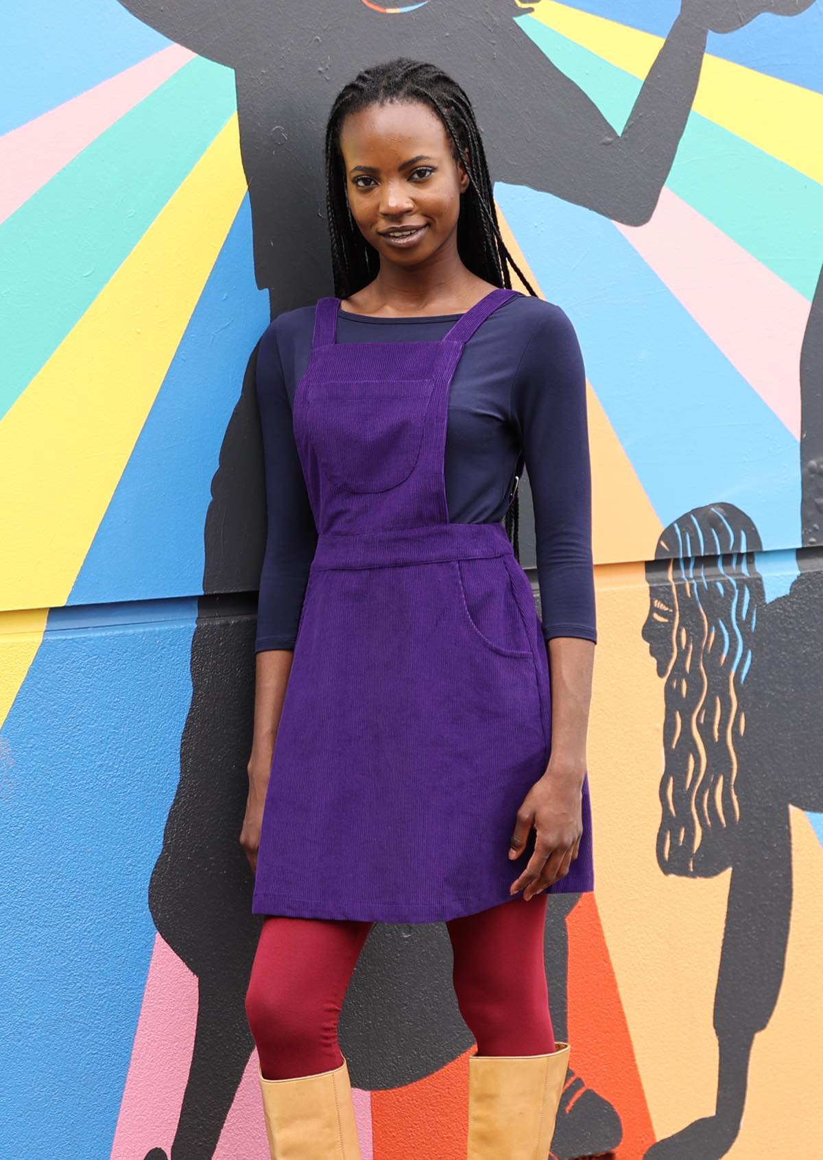 woman wearing purple cotton corduroy pinafore over navy blue top and maroon leggings  leaning against bright painted wall