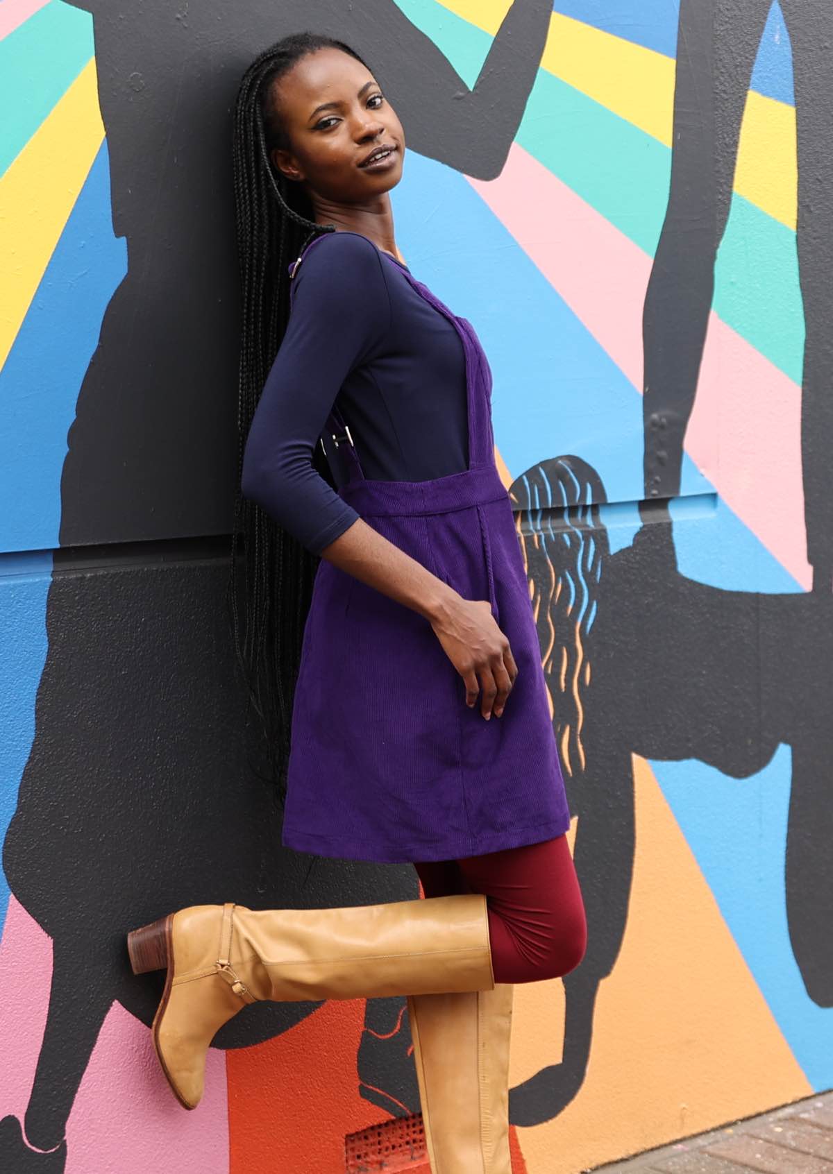 woman wearing purple cotton corduroy pinafore over navy blue top and maroon leggings  and long tan boots