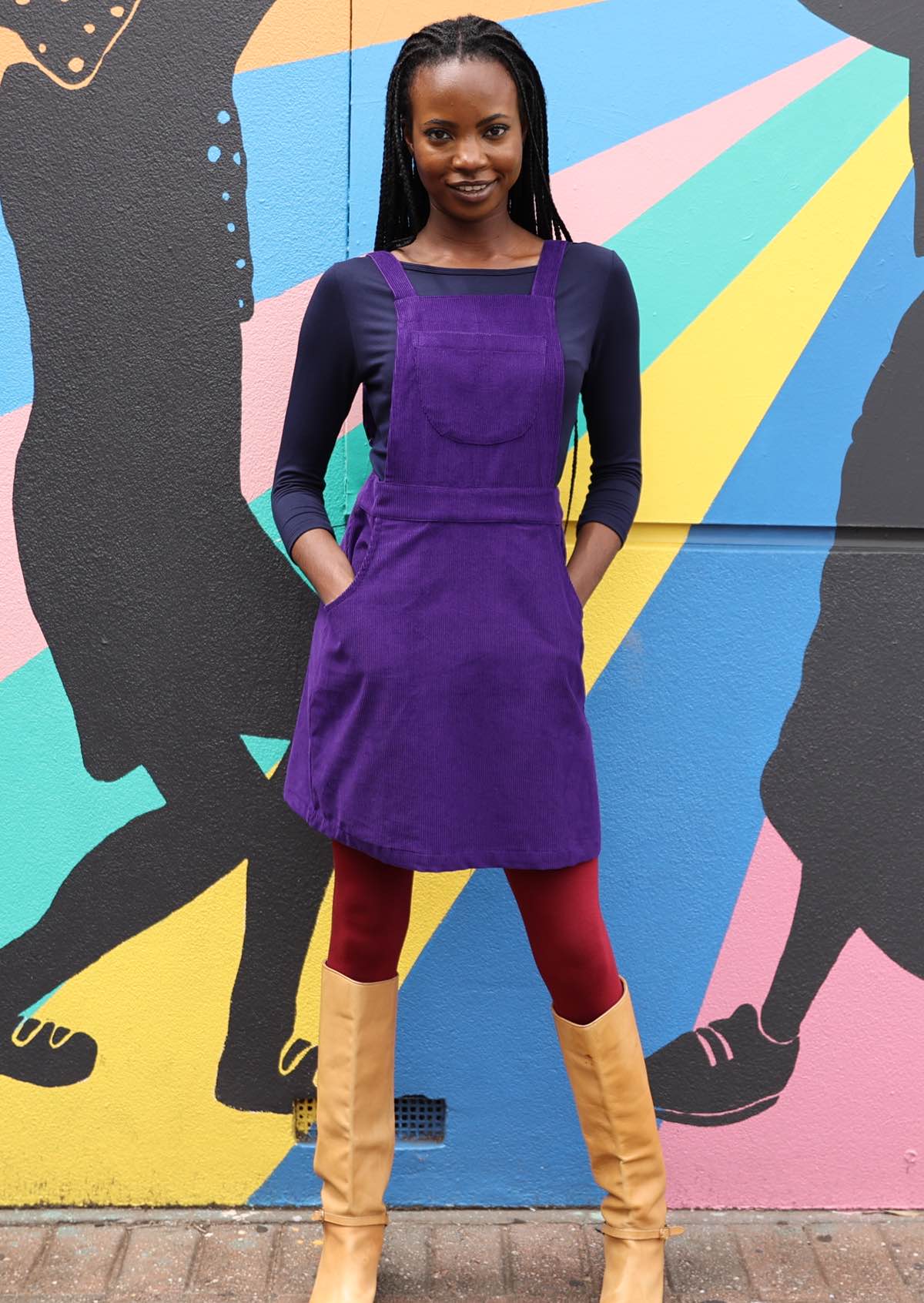 woman wearing purple cotton corduroy pinafore over navy blue top and maroon leggings with hands in pockets