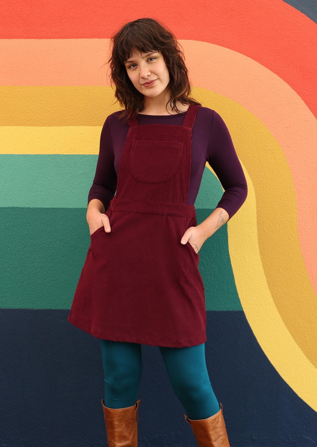 woman wearing maroon cotton corduroy pinafore over purple top and teal leggings  with hands in pockets