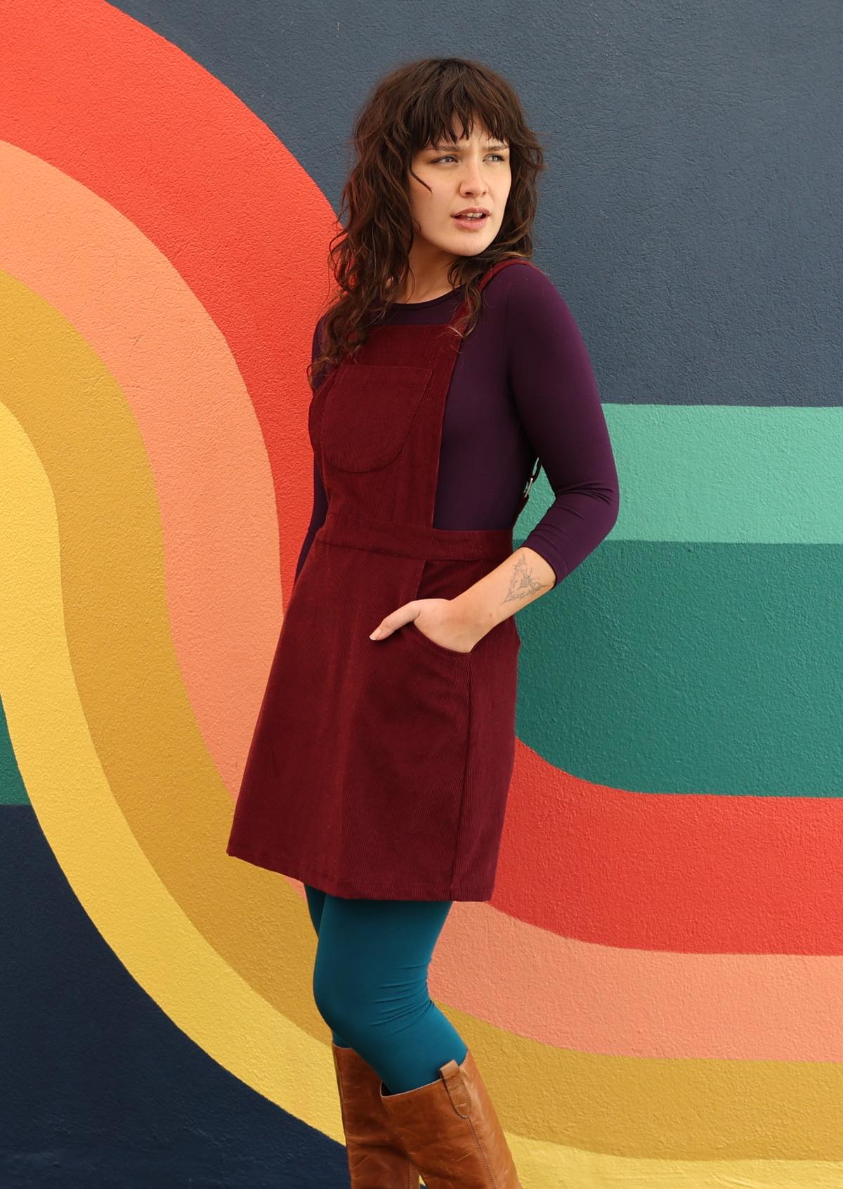 woman wearing maroon cotton corduroy pinafore over purple top and teal leggings  in front of 70's style mural