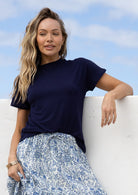 Woman wearing a navy t-shirt and blue floral skirt, leaning against a white wall