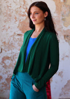 Woman stands side on in forest green long sleeve stretch rayon cardi with a waterfall front hem that is cropped at the front whilst back is longer