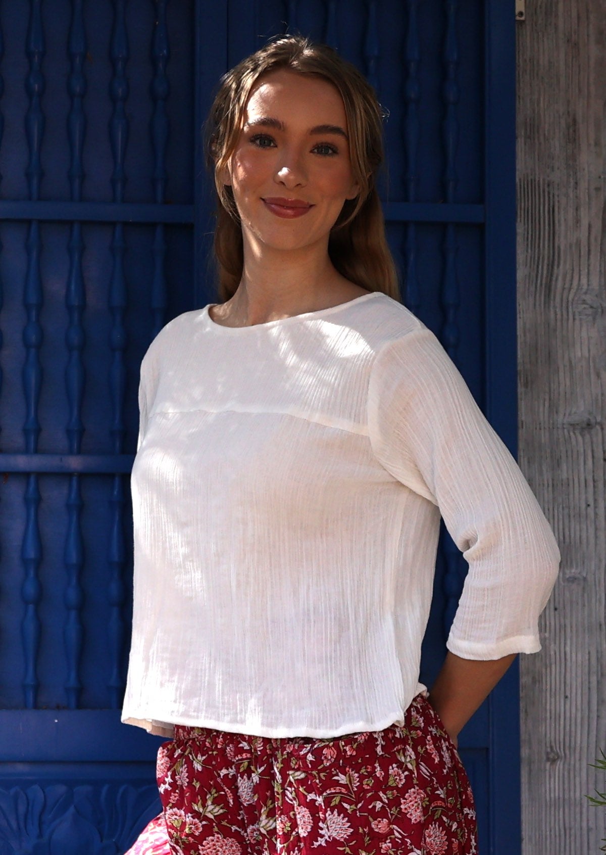 Woman wearing a white textured cotton top and red floral pants standing against a blue wall.