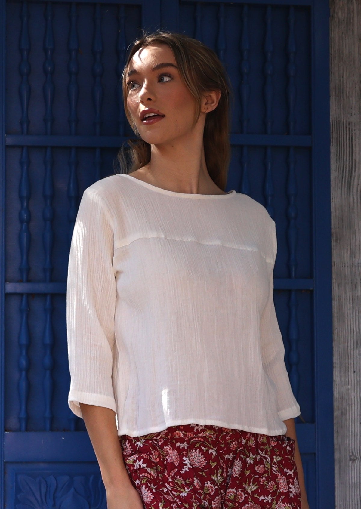 Woman wearing a white cotton top with floral pants against a blue wall.