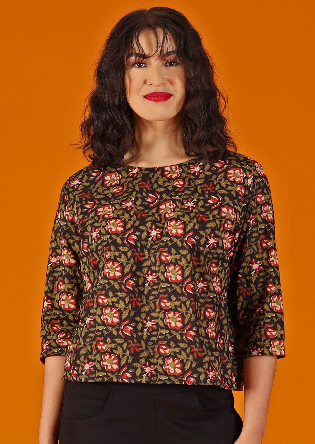 woman with dark hair and red lipstick in boxy cotton blouse, black with floral design  worn with black cotton pants