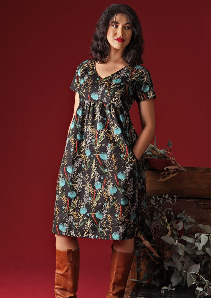 Model wears Etta Dress Thistle V-neck short sleeve below knee length cotton dress with empire waistline in botanical print with black base 