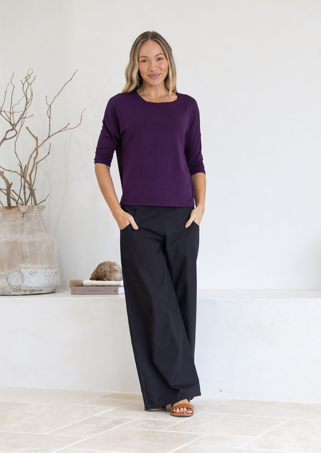 Woman wearing a purple relaxed fit top with 3/4 sleeves and black pants standing in a minimalistic room.