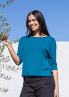 Woman wearing a teal relaxed fit top and black pants standing next to an olive tree