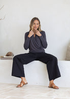 Woman sitting on a white ledge wearing a dark grey crossover long sleeve top and black pants.