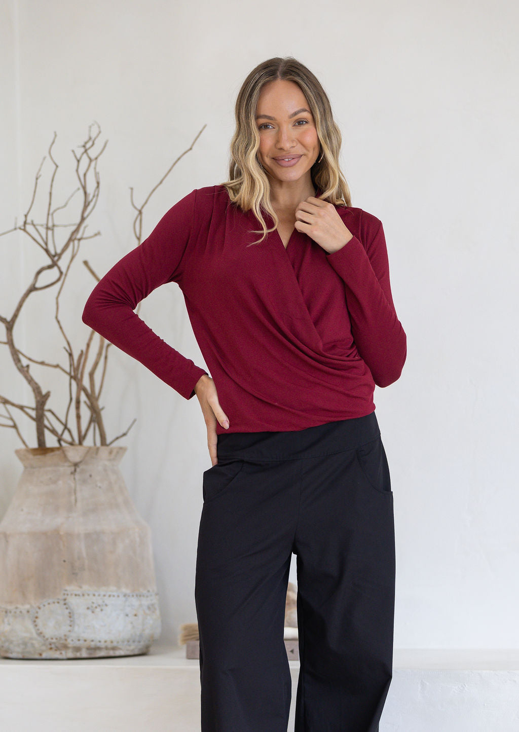 Woman wearing a maroon long sleeve crossover top with pleats at front of shoulders and black pants 