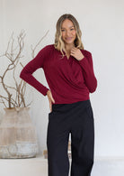 Woman wearing a maroon long sleeve crossover top with pleats at front of shoulders and black pants 