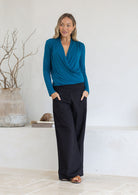 Woman wearing a teal crossover top with pleats at the front of the shoulders paired with black pants