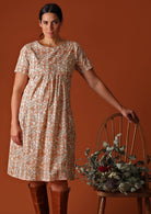 Model wears a 100% lightweight cotton knee length dress with pockets. The floral pattern of peach and olive green is on a cream base. This short sleeve dress is loose fit and lined.