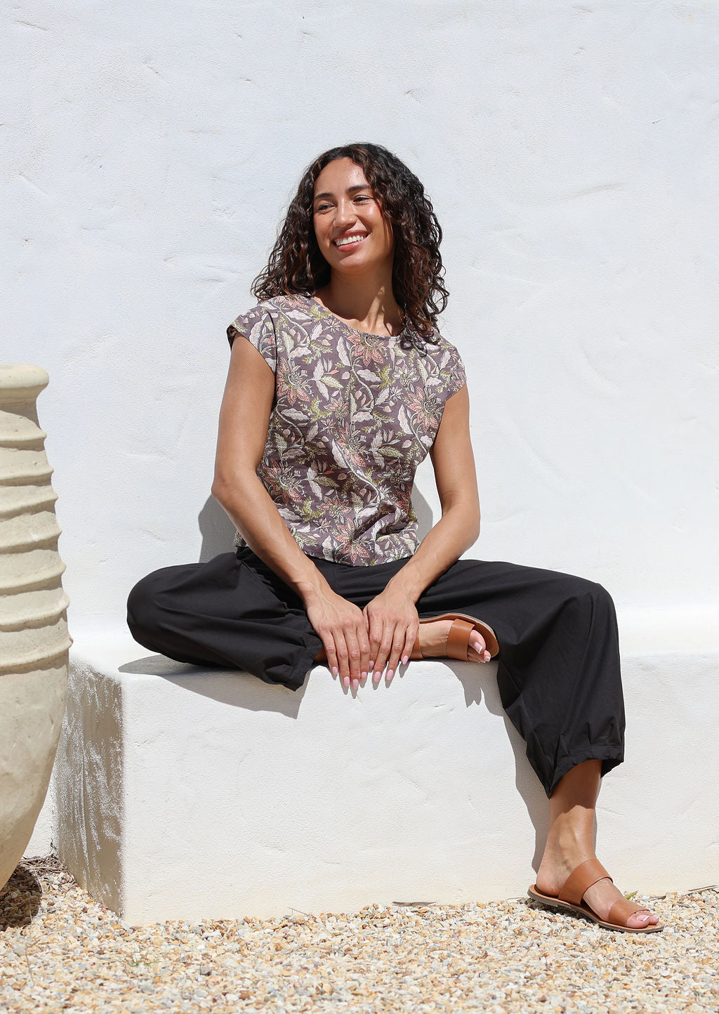 Woman sitting on a white concrete bench wears 100% cotton black pants with pleated ankles and pockets