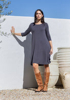 Woman wearing a long-sleeve grey jersey dress and brown boots standing against a white wall with blue sky.