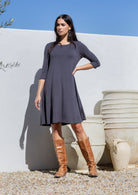 Woman wearing a dark gray stretch jersey dress and brown knee-high boots standing against a white wall with large pots.