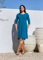 Woman wearing a teal blue jersey dress and brown boots standing against a white wall with decorative pots.