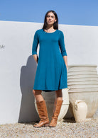 Woman wearing a  teal blue jersey dress and brown boots standing against a white wall with a blue sky.