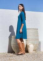 Woman in a teal stretch rayon dress and brown boots standing against a white wall with large beige pots.