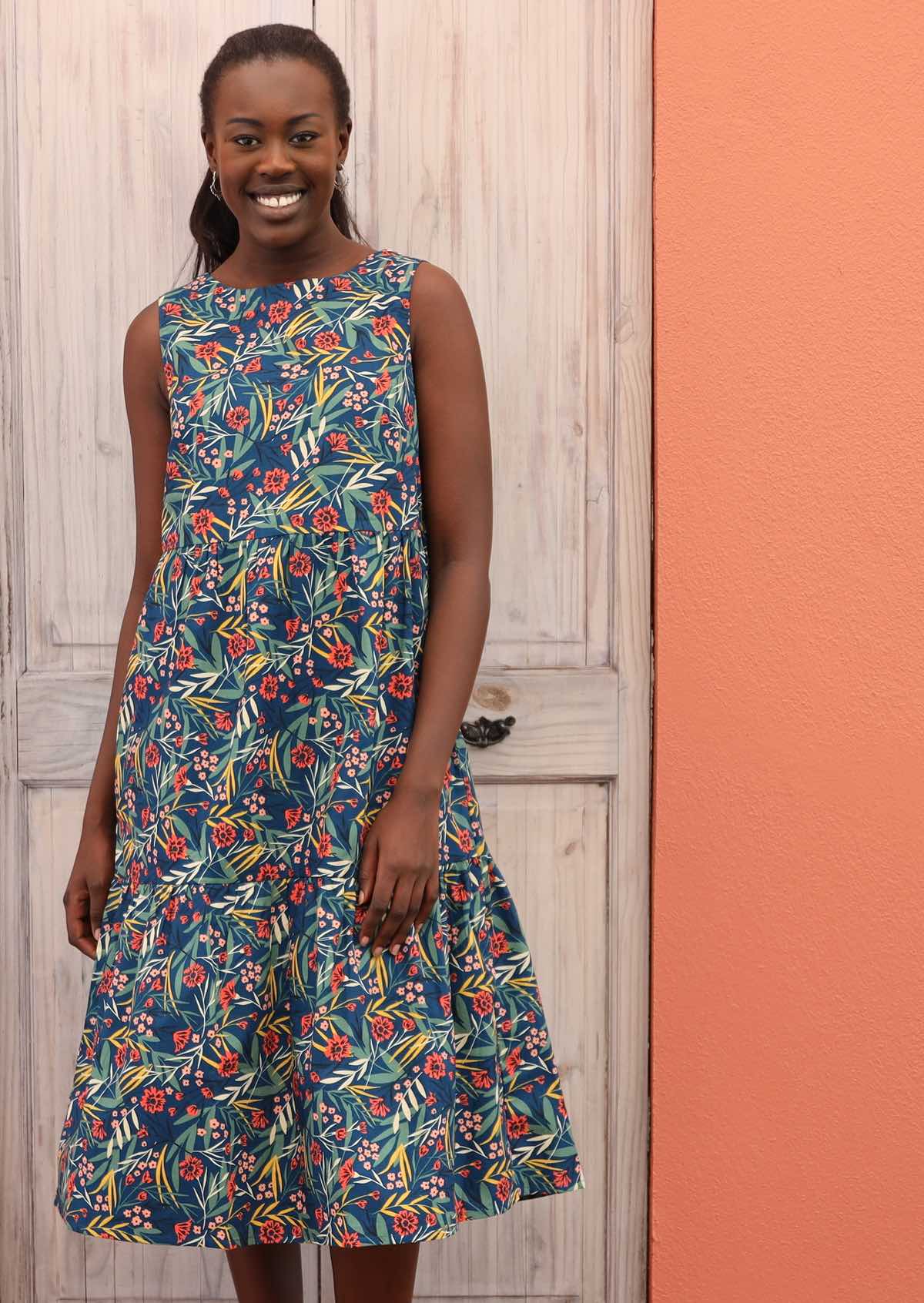 Smiling model wears a sleeveless, three tiered dress with a floral print on a blue base.